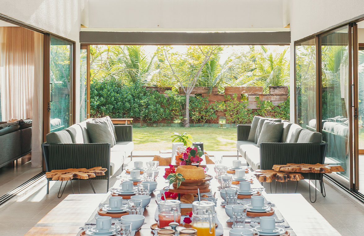 Mesa de café da manhã com bolos, sucos e flores na sala integrada com sofás e jardim ao fundo
