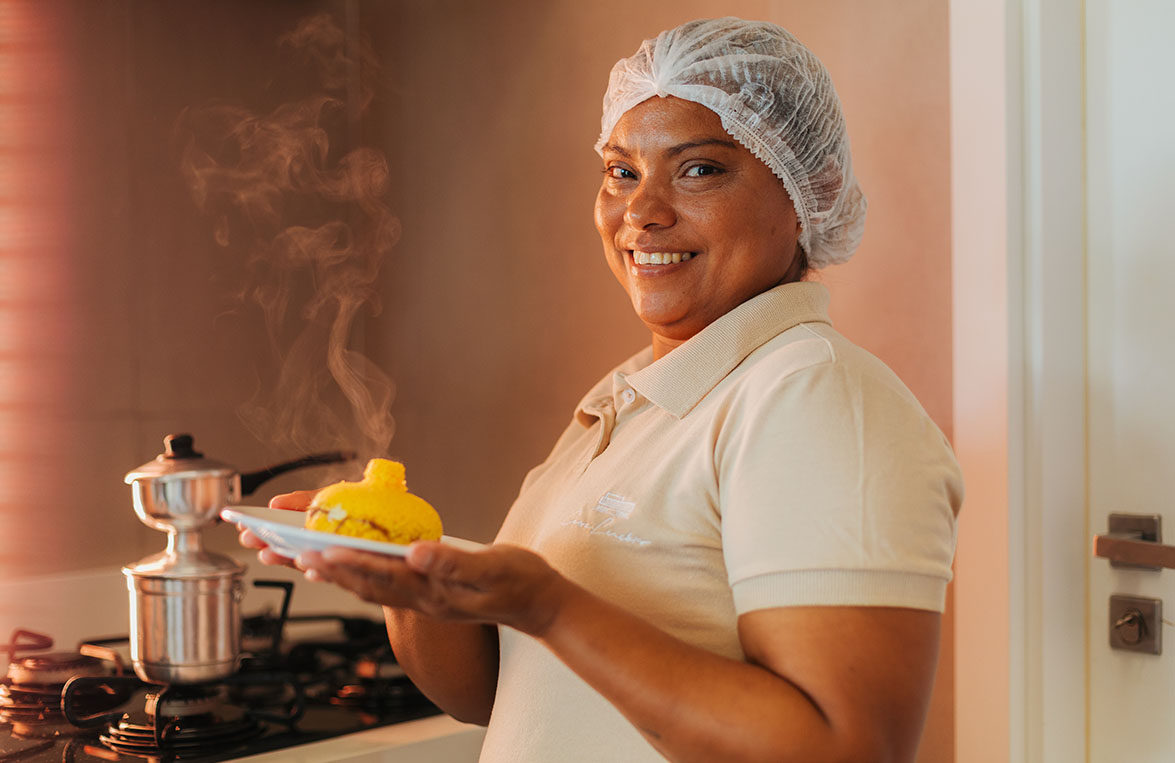 Cozinheira servindo cuscuz no prato ao lado do fogão com panela ao vapor