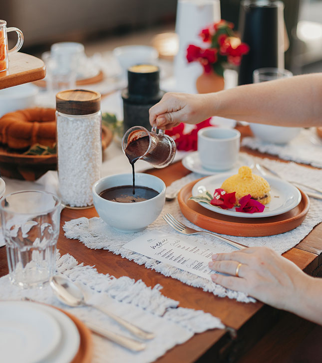 Mesa de café da manhã com chocolate quente sendo servido, bolo e flores