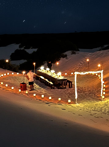 Jantar nas dunas iluminado por velas e lanternas sob o céu estrelado