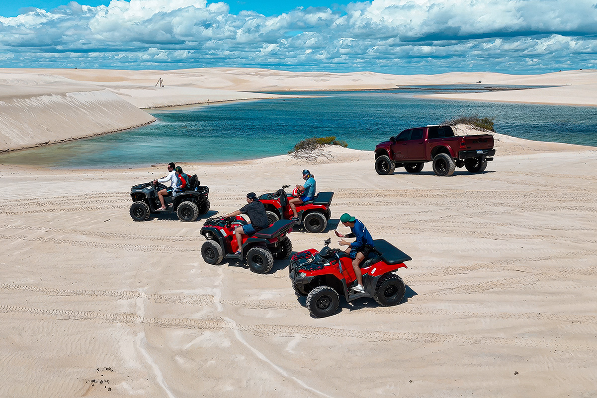 Turistas pilotando quadriciclos pelas dunas com lagoas e aerogeradores ao fundo