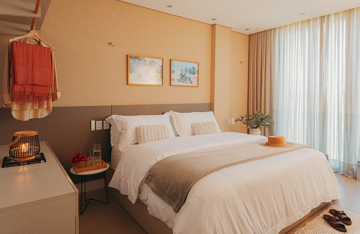 Suíte com cama king size, roupa de cama branca, quadros decorativos e cortina translúcida
