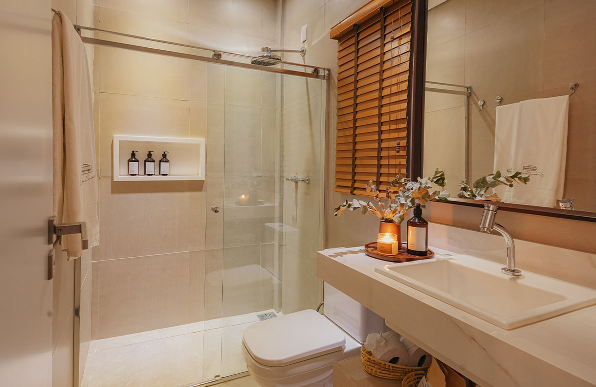 Banheiro da suíte com box de vidro, amenities, persiana de madeira e bancada com vela e flores